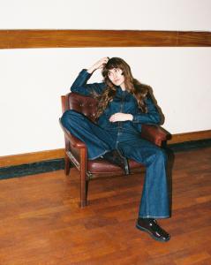 Personne assise dans un fauteuil en cuir, portant un ensemble en denim brut Nudie Jeans, dans un intérieur simple avec sol en bois.