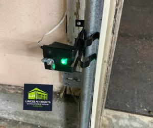 A close-up of a black garage door safety sensor with a glowing green light attached to a metal track, featuring the Lincoln Heights Garage Door Repair Experts logo.