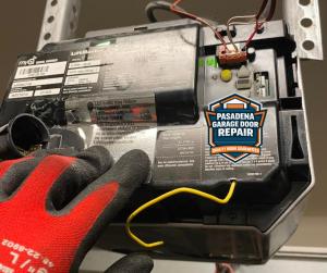 A technician in a red work glove inspecting the internal circuit board and wiring of a LiftMaster garage door opener, featuring the Pasadena Garage Door Repair logo.