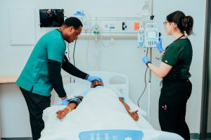 Photo shows two nursing students wearing green scrubs in a nursing simulation lab with a patient simulator in a hospital bed.