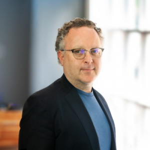 Headshot of Kevin Buerger, a man with glasses and short curly hair, wearing a dark blazer over a blue shirt, smiling in a professional office setting.