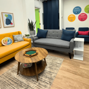 A cozy living space featuring a gray sofa, yellow couch, wooden coffee table, and colorful wall decor. Natural light streams in.