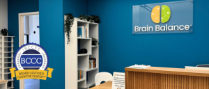 Interior of a Brain Balance center, featuring a blue wall, reception desk, bookshelf, and a certification badge for autism support.