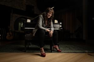 Singer Songwriter Author Nancy Manet sits on a piano bench, facing the camera with her back to the piano. it is a casual pose as if listening to the viewer.  she smiles. she is wearing a black and white checked blazer, black pants and dark red shoes.