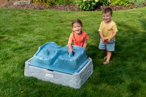 Two children playing with cars on the lid of a sandbox that looks like a mountain range.