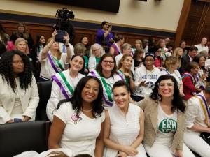 ERA Congressional Hearing, May 2019 - sitting left to right, Jennifer Carroll Foy, Alyssa Milano, Kate Kelly; a group of women sit behind them; everyone is wearing white, many are smiling