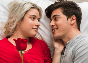 A couple lying together on white bedding, sharing an intimate Valentine's Day moment with a red rose, representing emotional connection and relationship wellness during the holiday season.