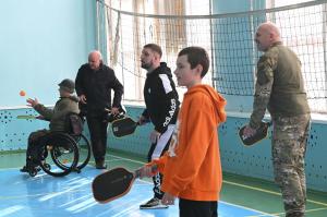 Four people playing pickleball indoors on a blue court, including a man in a wheelchair, a man in military fatigues hitting the ball, a civilian in athletic wear, and a young boy in an orange hoodie.