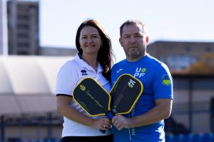 Anna Glotova and Volodymyr Svirskyi standing side-by-side outdoors on a sunny day, both holding black and yellow Anywhere Pickleball paddles. Volodymyr is wearing a blue athletic shirt featuring the UPF logo.