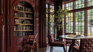 Luxurious home library interior in rich burgundy tones with wooden arched bookshelves filled with colorful print books, potted plant, round table with chessboard, upholstered chairs, and large arched windows overlooking greenery, as showcased by MYTSV.com