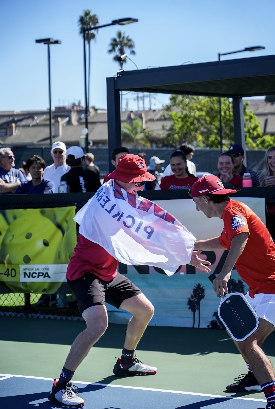 Two collegiate pickleball teammates celebrating on court during an NCPA championship match, with spectators cheering in the background