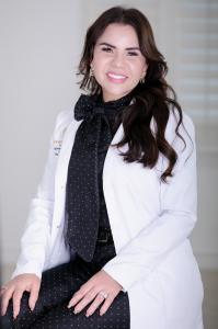 Portrait of Dr. Barbara Ortiz wearing a white medical coat and black polka-dot blouse, smiling in a professional office setting at Your Dream Aesthetics.