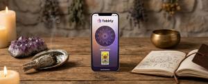 Ahşap masada spiritüel objeler arasında duran ve Tabirly mobil uyumlu web sitesindeki astroloji çarkı ile Tarot 'Günün Kartı' özelliğini gösteren akıllı telefon ekranı.