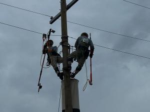 Utility linemen wearing flame-resistant clothing and safety harnesses climb a power pole to service overhead electrical lines.