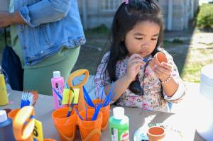 A young child carefully paints a small clay pot at an outdoor STEM activity table, surrounded by paint bottles, brushes and craft supplies during a GSSM community event.