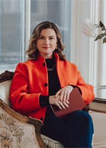 Manja Horner, Author of Pass The Torch - A Rallying Call to Rescue The Future of Trades, holding a book while sitting on a couch in an orange suit jacket