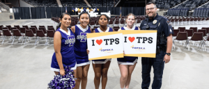 A group of cheerleaders in uniforms and a police officer hold signs reading "I ❤️ TPS" in an arena with empty seats behind them.