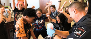 A group of young people with raised hands surrounds a police officer demonstrating a science experiment involving colorful liquids.