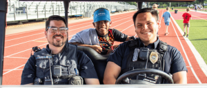 Two police officers sit in a golf cart while a child stands behind them, smiling. A track and field area is visible in the background.