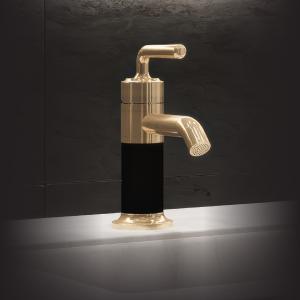 A modern, gold-colored bathroom faucet with a black cylindrical base stands on a white marble countertop against a dark tiled wall. The faucet has a curved spout with a perforated nozzle and a matching gold handle on top, illuminated by soft, dramatic lig