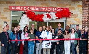 A group of Ally Medical Emergency Room physicians, staff, and community members stand together holding a red ribbon in front of the Burleson facility entrance, decorated with red and white balloons, during the official grand opening ribbon cutting ceremon
