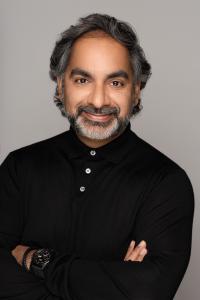 Photograph of Doctor Samir Qamar, dressed in a black knit shirt, buttoned up, arms folded and smiling.