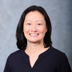 Woman with straight black hair wearing a dark top, smiling in front of a light gray background.