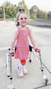A young girl with CP stands smiling broadly in her walker and orthotics in a red and white striped dress and pink glasses.