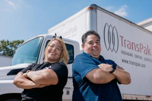 Two TransPak employees stand back to back with arms crossed, smiling confidently in front of a TransPak branded delivery truck on a sunny day.