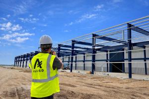 Construction worker wearing a white hard hat and a bright yellow ALLIED safety vest photographing a large blue steel building under construction, with white metal wall panels and a clear blue sky overhead.