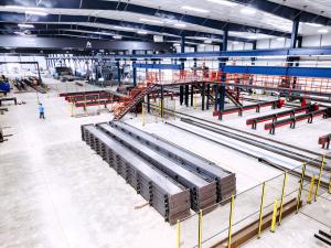 Wide-angle view of a modern steel fabrication facility with organized stacks of structural steel beams, automated processing lines, overhead cranes, and a red elevated work platform inside a brightly lit industrial building.