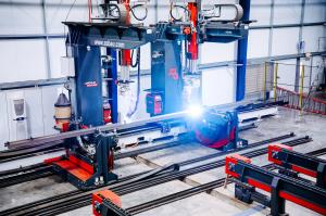 Automated structural steel processing line inside a modern fabrication facility, featuring robotic welding equipment, precision rails, and a bright active weld point on a steel member.