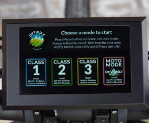 Close-up of the Optibike G2 Altus handlebar-mounted MMI display showing selectable riding modes including Class 1, Class 2, Class 3, and High-Power Off-Road Moto Mode with wattage and speed limits.