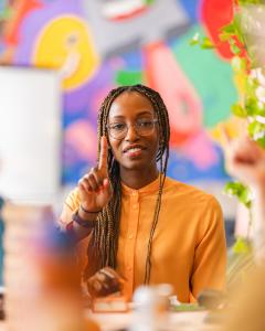Nile Livingston, CEO and Founder of Creative Repute, wearing an orange blouse and glasses, gestures while leading a team discussion in front of a colorful mural background.