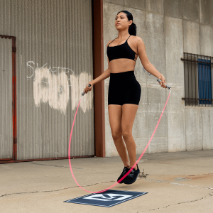 Athletic woman jumping rope outdoors using the Rx Jump Rope 2.0 with a pink cable, wearing black workout apparel and mid-air during a speed training session.