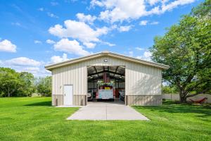 Front view of a completed pre-engineered steel building with an open garage door, concrete driveway, and vehicles visible inside, set on a grassy property with trees and blue sky.