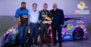 Young white male racercar driver in a black race suit flanked by three older men posing for a group photo in front of his racecar