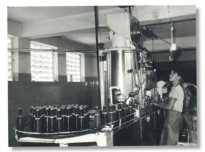 Black-and-white photo of a 1970s bottling line in Munich producing Paulaner cola-orange beverage bottles.