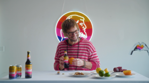 Person holding a Paulaner Sunset bottle at a table with citrus fruits and cans, colorful logo sign in the background.