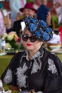 Woman seated at a Kentucky Derby themed event wearing a dramatic black fascinator adorned with blue floral appliqués and pearls. She pairs the hat with oversized black sunglasses, red lipstick, pearl earrings, and a sheer black blouse with white embroider