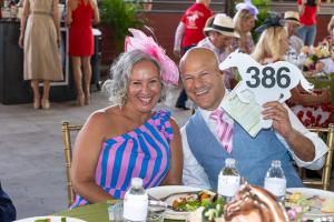 Smiling couple seated at a Kentucky Derby themed event. The woman wears a pink fascinator and a pink and blue striped one shoulder dress, while the man wears a light blue vest with a pink tie and holds a white horse shaped auction paddle labeled “386.” Pl