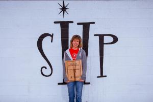Susie Asadorian stands alone in front of a large Sleep in Heavenly Peace logo painted in black on a white cinder block exterior wall. She smiles at the camera, holding a wooden engraved Guardian of the Mission award plaque with both hands. She wears a gra