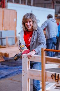 Susie Asadorian smiles warmly at the camera as she uses a yellow and black DeWalt orbital sander on a light wood bed frame inside an SHP workshop. She wears a gray zip-up hoodie over a red SHP OH-Cuyahoga t-shirt and blue jeans. The SHP logo is branded in