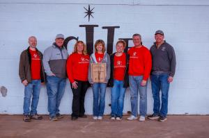 A group of seven adults poses in a row in front of the Sleep in Heavenly Peace logo painted on a white cinder block exterior wall. Susie Asadorian stands in the center holding the wooden Guardian of the Mission award plaque. Five of the seven wear red SHP