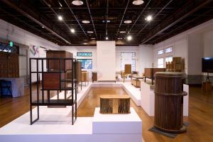 An interior view of a historic exhibition hall with wooden floors and exposed ceiling beams, displaying modern wooden furniture pieces and cabinets on white geometric pedestals for the Daxi Wood Furniture Craft Design Competition.