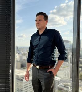 Photo of Piotr Siwek, Global Center of Excellence Manager at Mitsubishi Electric Factory Automation Global, standing in a modern office overlooking a city skyline, representing leadership in industrial automation and digital transformation.