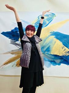 A happy artist/woman with her arms raised in front of a gold and blue, abstract painting