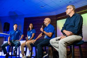 Four Allied Steel Buildings executives, Chris Rucker, Catherine Soto, Michael Lassner, and Sergio Plaza, seated on stage with microphones, speaking to employees during a company event under blue stage lighting.