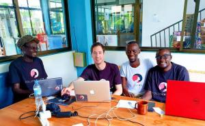 Photo of the Follow Alice founding team during their first meeting in Tanzania in 2014. From left to right: Chris, Reto, Kazi, and Robert sit at a wooden desk with laptops. They are all smiling, wearing Follow Alice branded t-shirts, marking the beginning