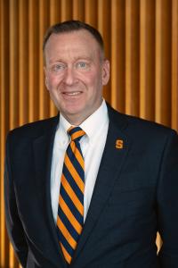 Image of Syracuse University Chancellor-elect J. Michael Haynie wearing a dark blue suit jacket, white button-down shirt, an orange and blue tie